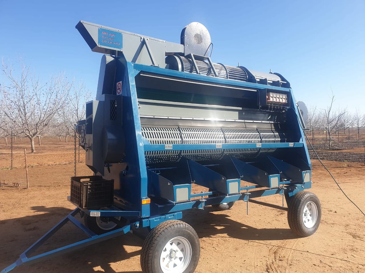 Blue SMJ pre-cleaner on wheels with visible screens and collection outlets in an orchard